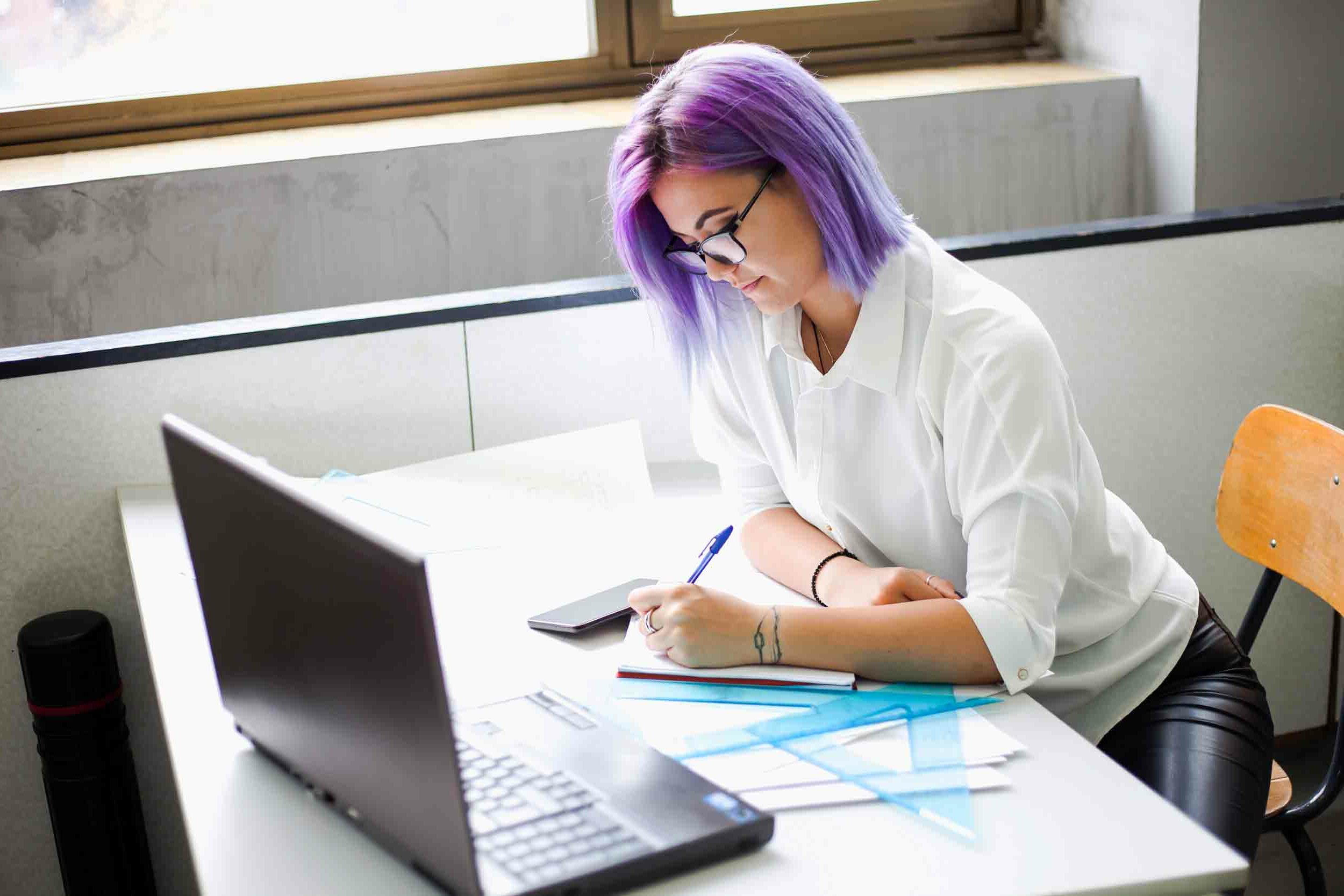 Young women with purple hair watching something on laptop and writing something down. She is working on some project