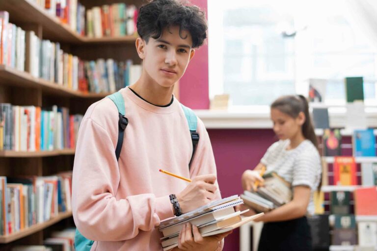 Clever teenager in sweatshirt looking at you while standing by bookshelf in college library and making notes in notepad