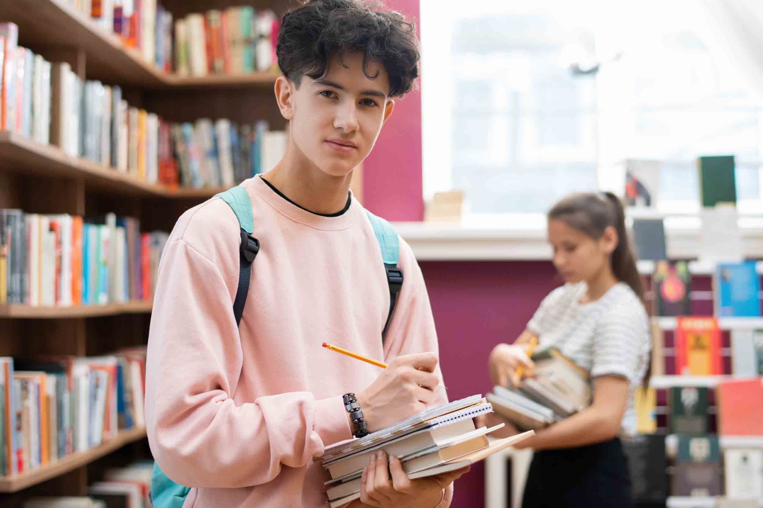 Clever teenager in sweatshirt looking at you while standing by bookshelf in college library and making notes in notepad